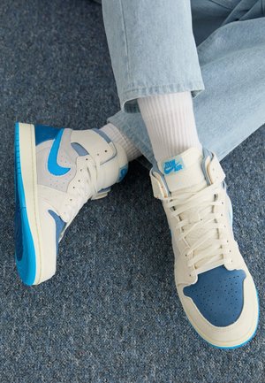 Personne portant des baskets Nike Zoom Air blanches et bleues, des chaussettes blanches et un jean bleu clair, assise en tailleur sur une moquette bleue.