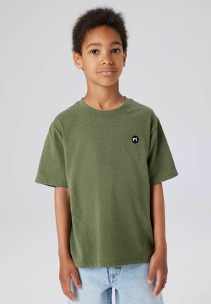 Young boy with short curly hair wearing a green t-shirt and light blue jeans, standing against a plain light background.