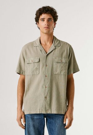 Giovane uomo con capelli ricci che indossa una camicia beige a maniche corte con bottoni e due tasche sul petto, abbinata a jeans blu, in piedi di fronte a uno sfondo semplice.