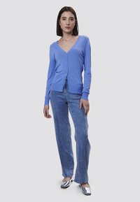 Cardigan azzurro chiaro con scollo a V e chiusura con bottoni, abbinato a jeans in denim chiaro con vita alta. Il modello si trova su uno sfondo neutro.