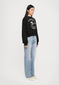 Mulher de pé vestindo um moletom preto com texto e gráfico brancos, jeans azul claro com estampado floral em rosa e sapatos de salto agudo brancos.