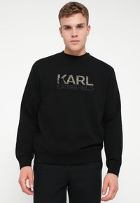 Mladý muž oblečený v čiernej mikine Karl Lagerfeld a čiernych nohaviciach, stojaci pred jednoduchým bielym pozadím.