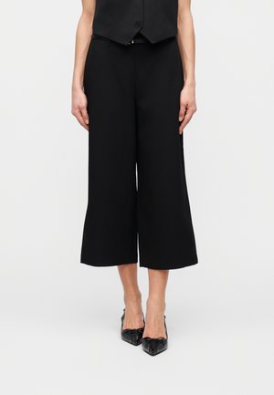 Frau trägt schwarze, weit geschnittene, verkürzte Hosen, schwarze spitze Absatzschuhe mit Schleifen und eine schwarze ärmellose Weste, steht vor einem weißen Hintergrund.