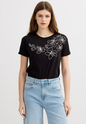 Mujer joven con camiseta negra de manga corta con diseño floral en línea blanca y jeans de tiro alto azul claro de pie frente a fondo blanco.