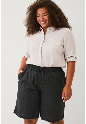 Vrouw met krullend haar die glimlacht, draagt een lichtbeige overhemd met knopen en zwarte korte broek met trekkoord, met handen in de zakken.