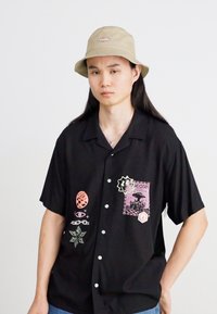 Chemise noire à manches courtes avec divers écussons graphiques, fermeture à boutons et coupe décontractée, accompagnée d'un chapeau seau beige.