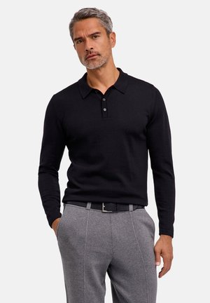 Man met grijs haar met een zwart poloshirt met lange mouwen, ingestopt in een grijze broek met een zwarte riem, staat met één hand in de zak.