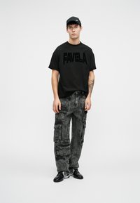 Černé tričko s grafikou "FAVELA", šedé cargo kalhoty s kapsami a černá obuv. Oblečení je ve volném, oversized střihu.