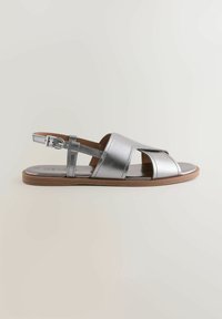 Sandales métalliques argentées avec design à brides croisées, boucle de cheville ajustable, semelle marron et texture lisse. Bout ouvert avec des détails minimalistes.