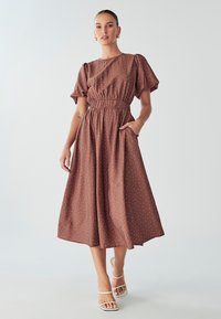 Smeđa midi haljina s točkicama, kratkim puf rukavima i elastičnom strukom, s džepovima i lepršavom suknjom. Nosena sa bijelim sandalama.