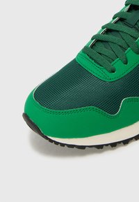 Grønn sneakers med meshupperside og syntetiske overlay, rund tå, teksturerte lisser, samt en kontrasterende hvit mellomsåle og svart yttersåle.