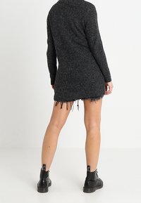 Czarna, teksturowana sukienka swetrowa z długimi rękawami, z postrzępionym dołem i dopasowanym krojem. Noszona z czarnymi botkami za kostkę.
