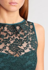 Taurfarbene Spitzenkleid mit floralen Mustern und einem durchsichtigen Ausschnitt, der mit einem einfarbigen taubfarbenen Saum veredelt ist. Enganliegendes Design mit glatter Textur.