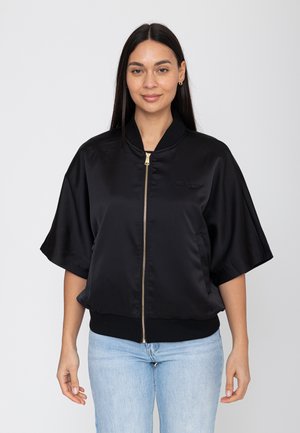 Kobieta z długimi ciemnymi włosami, ubrana w czarną krótko rękawową kurtkę z zamkiem i jasnoniebieskie dżinsy, stojąca na tle jednolitego jasnego tła.