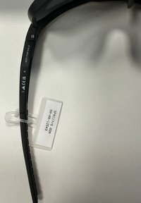 Braccio della montatura degli occhiali neri con la dicitura "NON IN VENDITA" e marchi di certificazione, attaccato a un'etichetta di plastica con codici prodotto su una superficie bianca.