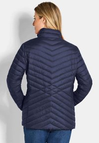 Marineblauwe pufferjas met een chevron-quiltpatroon, hoge kraag, lange mouwen en een zachte textuur, gecombineerd met blauwe spijkerbroek.