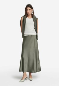 Olivengrüner Satin-Maxi-Rock kombiniert mit einem weißen gerippten Tanktop und einer grauen ärmellosen Strickjacke. Weiße Slingback-Absätze mit strukturierten Akzenten.