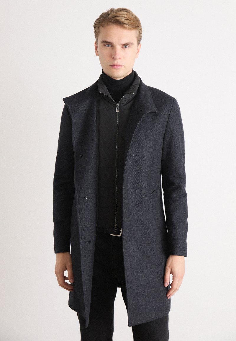 Manteau en mélange de laine gris foncé à coupe ajustée, col pointu et fermeture à boutons ; superposé à une veste intérieure noire matelassée à zip.