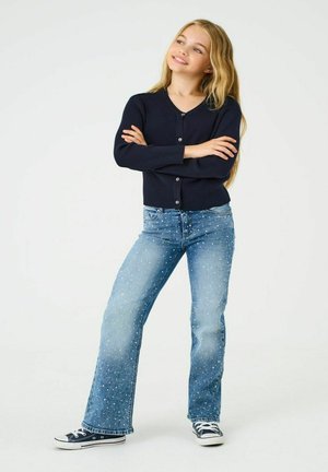 Námořnický žebrovaný svetr na knoflíky; světle modré džínové kalhoty s rozptýlenými bílými skvrnami; černé plátěné tenisky s bílou podrážkou a tkaničkami.