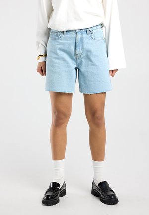 Hellblaue Jeansshorts, getragen mit weißen Socken und schwarzen Loafers, kombiniert mit einer langärmeligen weißen Bluse vor neutralem Hintergrund.