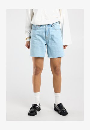 Hellblaue Jeansshorts, getragen mit weißen Socken und schwarzen Loafers, kombiniert mit einer langärmeligen weißen Bluse vor neutralem Hintergrund.