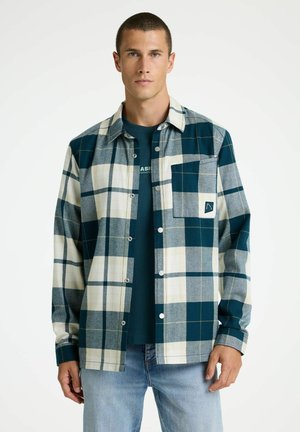 Muž oblečený v modrej, sivej a bielej kockovanej košeli na gombíky, cez ktorú má tmavomodré tričko a svetlomodré rifle, stojí pred jednoduchým pozadím.