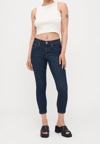 Jean bleu foncé à coupe slim, doté d'un design à cinq poches et de coutures légères. Associé à un crop top blanc ajusté et des sandales à plateforme noires.
