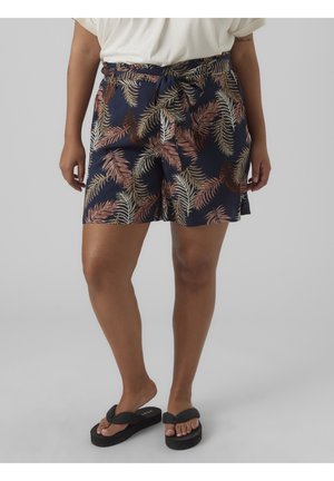 Mujer lleva shorts azul marino con patrones de hojas marrones y beige, combinados con chanclas negras y una camiseta blanca sobre un fondo gris liso.
