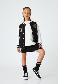 Giacca varsity bianca e nera con ricamo "Whatever 86", abbinata a un maglione bianco, una gonna nera e calze fino al ginocchio nere con strisce.