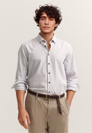 Junger Mann mit lockigem Haar, der ein weißes, langärmliges, gestreiftes Hemd mit hochgekrempelten Ärmeln, beigen Hosen und einem braunen geflochtenen Gürtel trägt.