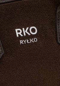 Izbliza smeđi suede materijal s bijelim logotipom "RKO RYŁKO" i tamnosmeđim kožnim rubom prošivenim na ivici.