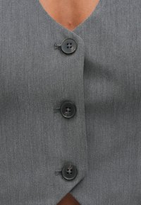 Sivá vesta s hlbokým výstrihom do V, vybavená tromi čiernymi gombíkmi a textúrovanou látkou. Významné detaily zahŕňajú prešívanie a zaoblený spodný okraj.