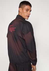 Melnā un bordo krāsas sporta jaka ar izšūtu krokodila logo aizmugurē, elastīgām aprocēm un gludu, vieglu tekstūru.