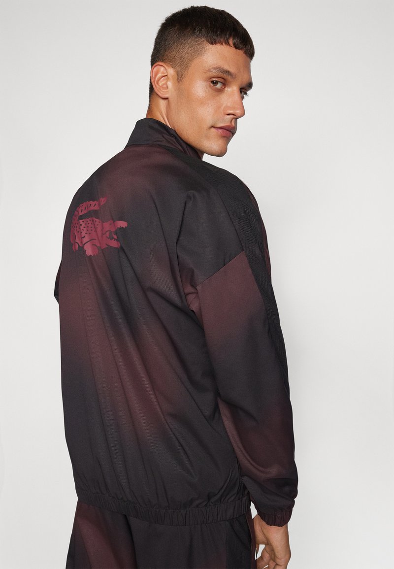 Schwarze und burgunderfarbene Trackjacke mit einem gestickten Krokodil-Logo auf dem Rücken, elastischen Bündchen und einer glatten, leichten Textur.