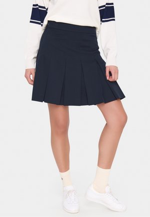 Femme portant une jupe plissée bleu marine, des baskets blanches, des chaussettes crème et un pull blanc avec des rayures bleu marine aux manches, debout sur un fond blanc.