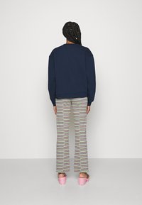 Person med flätat hår som bär en marinblå sweatshirt, flerfärgade randiga byxor och rosa platåsandaler som står mot en vit vägg.