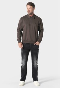 Felpa marrone con zip e colletto, abbinata a jeans neri sbiaditi e sneakers bianche. Cuciture e logo distintivi sul petto.