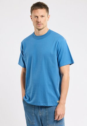 Homme portant un t-shirt bleu uni à manches courtes et un pantalon en denim, debout avec une main dans la poche devant un fond blanc uni.