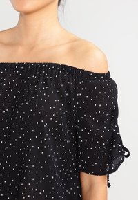 Schwarzes Off-Shoulder-Top mit weißen Punkten, aus strukturiertem Stoff, mit elastischem Ausschnitt und lockeren, kurzen Ärmeln mit Bindebändern.