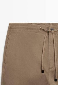 Beżowe spodnie z elastycznym pasem i sznurkiem do wiązania, wykonane z gładkiego, teksturowanego materiału z subtelnymi diagonalnymi wzorami.