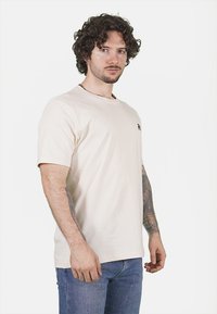 T-shirt in cotone beige con maniche corte, colletto a girocollo e un piccolo logo nero sul lato sinistro del petto. Il modello indossa jeans blu.