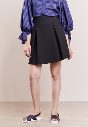 Frau trägt einen schwarzen plissierten Minirock, eine violette Satinbluse mit Puffärmeln, die an den Bündchen gebunden sind, und schwarze Sandalen mit weißen Sohlen.