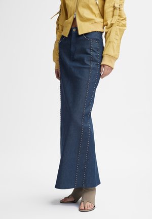 Blå denim maxi nederdel med nitte detaljer i siden, båret med brune åbne hæle støvler og en kort gul jakke.