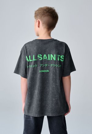 Chłopiec tyłem ubrany w czarną koszulkę All Saints z zielonym napisem, zawierającym Tokyo, Underground i London na plecach.