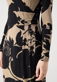 Mekko, jossa on musta ja beige väriyhdistelmä, pitkät hihat, kukkakuosi ja muotoiltu yläosa, jossa on kääntömuotoilu. Pehmeä, joustava kangas, jossa on sileä rakenne.