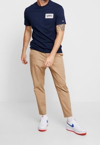 Navy cotton t-shirt with a rectangular white logo reading "TOMMY JEANS" on the chest, paired with beige chinos and white sneakers with blue accents.