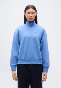 Ανοιχτό μπλε φλις φούτερ με ψηλή λαιμόκοψη, κλείσιμο με φερμουάρ μέχρι τη μέση και ριμπ μανσέτες· διαθέτει μικρό πράσινο λογότυπο στο στήθος.