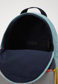 Zaino azzurro chiaro con interno blu navy, dotato di una tasca con zipper, materiale liscio e maniglia superiore. Include un tirazip giallo brillante e nero.