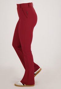 Slim-fit donkerrood broek gedragen met witte sneakers met beige en zwarte accenten, weergegeven vanaf de zijkant op een neutrale achtergrond.