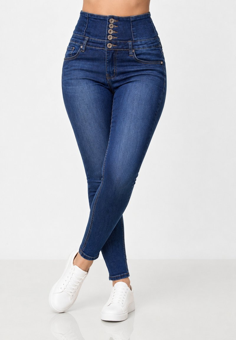Kvinde iført højttaljede blå skinny jeans med fem knapper og hvide sneakers, stående med benene krydset på en hvid gulv.